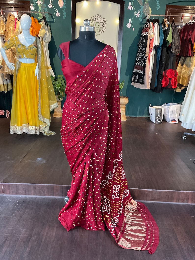 May include: A burgundy saree with a dotted pattern and a decorative border is draped on a mannequin. The saree has a deep V-neckline and is displayed in a boutique setting. Other garments are visible in the background.
