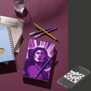 May include: A purple notebook with a woman's face on the cover. The woman has long, dark hair and is wearing a purple dress. The notebook is open to a blank page. There are pencils and pens on the table next to the notebook.