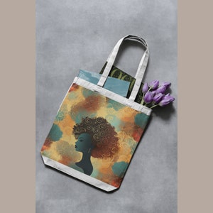May include: A white tote bag with a design of a woman with an afro hairstyle. The design is in shades of brown, yellow, and blue. The bag is lying flat on a gray surface.
