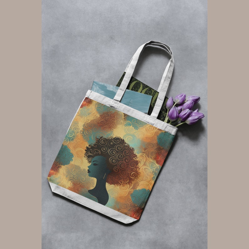 May include: A white tote bag with a design of a woman with an afro hairstyle. The design is in shades of brown, yellow, and blue. The bag is lying flat on a gray surface.