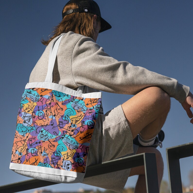 May include: A white tote bag with a colorful design of musical notes and abstract shapes. The bag is being held by a person wearing a grey sweatshirt and shorts.