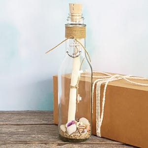 May include: A clear glass bottle with a cork stopper containing a rolled parchment scroll, sand, and small seashells at the base. The neck of the bottle is wrapped in tan twine with a silver-toned metal anchor charm tied to the front.