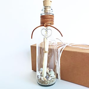 May include: A glass bottle with a cork stopper, filled with sand, seashells, and a rolled up piece of paper. The bottle is tied with brown leather cord and a silver heart charm with the number "3" on it.