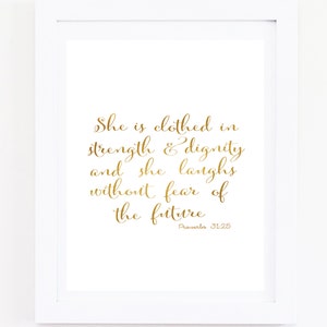 May include: Gold foil print of a Bible verse on a white background. The verse reads "She is clothed in strength & dignity and she laughs without fear of the future. Proverbs 31:25"