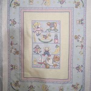 May include: A light blue and cream-colored baby quilt with a border of repeating cartoon characters and toys. The center features a rectangular panel with illustrations of a girl, a bear, a rocking horse, and other baby-themed images.