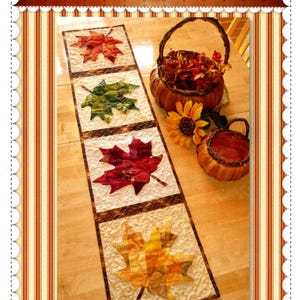 May include: A rectangular table runner featuring four square panels, each displaying a patchwork maple leaf in shades of red, green, and yellow. The background of each panel has a cream-colored quilted texture with a brown border.