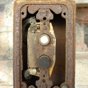Könnte beinhalten: Nahaufnahme eines verwitterten, rechteckigen Elektrokastens mit rostiger, brauner Oberfläche. Die Box hat eine kreisförmige Oberseite und zwei Knöpfe. Das Innere zeigt die Verkabelung und einen Schalter. Das Bild ist vor einer Backsteinmauer aufgenommen.