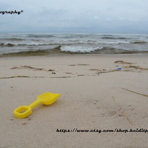 May include: A yellow plastic shovel rests on a sandy beach with the ocean in the background under a cloudy sky. The image includes the text "heiditphotography" and a website address.