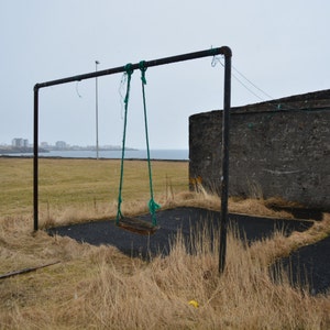 May include: A rusty swing set with a green rope swing hanging from it. The swing set is in a grassy field with a concrete wall in the background.