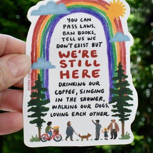 May include: A sticker with a rainbow and clouds, and the text "You can pass laws, ban books, tell us we don't exist but we're still here drinking our coffee, singing in the shower, walking our dogs, loving each other."