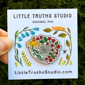 May include: White enamel pin with a spiderweb design and the text "It's all connected" in a circle around the web. The pin is surrounded by red, yellow, and green flowers. The pin is on a white card with the text "Little Truths Studio" and "LittleTruthsStudio.com".