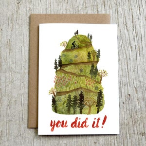 May include: A watercolor illustration of a mountain with a winding path and a person riding a bicycle up the hill. The text "you did it!" is written in red at the bottom of the card.