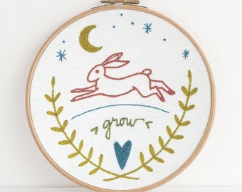Könnte beinhalten: Stickerei auf einem Reifen mit einem roten Hasen, der mit einem grünen Blätterkranz springt und dem Wort "grow" in grüner Schrift. Der Hintergrund ist weiß mit einem blauen Halbmond und Sternen.