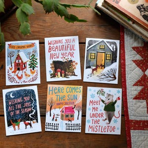 May include: Set of six illustrated holiday greeting cards. Designs include a red barn, a bear, a house with a dog, a winter scene with a dog, a house with the text "Here Comes The Sun", and a possum under mistletoe. Text includes "Cozy Christmas Wishes" and "Meet Me Under The Mistletoe".
