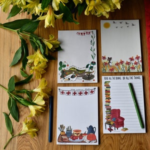 May include: Four different illustrated notepad designs. The first notepad features a white cat sleeping on a log with the text "Everything in its own time". The second notepad has a red, white, and blue bunting with the text "Happy sunshine days". The third notepad has a mouse sitting in a field of red flowers with the text "Read all the books, do all the things". The fourth notepad has a stack of books, a red armchair, and a cup of tea with the text "Read all the books, do all the things".