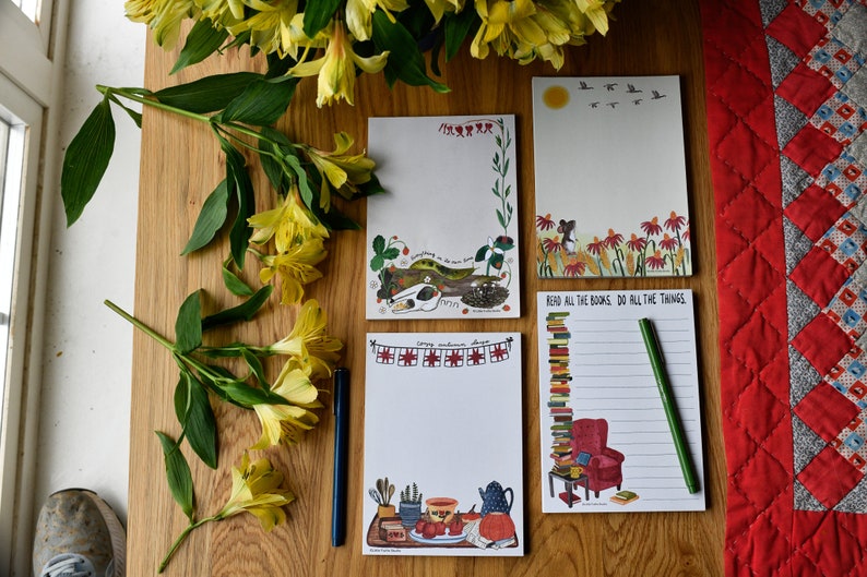 May include: Four different illustrated notepad designs. The first notepad features a white cat sleeping on a log with the text "Everything in its own time". The second notepad has a red, white, and blue bunting with the text "Happy sunshine days". The third notepad has a mouse sitting in a field of red flowers with the text "Read all the books, do all the things". The fourth notepad has a stack of books, a red armchair, and a cup of tea with the text "Read all the books, do all the things".