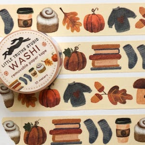 May include: A roll of Little Truths Studio washi tape with a fall theme. The tape features illustrations of pumpkins, books, sweaters, socks, coffee cups, cinnamon rolls, and leaves. The tape is 10 meters long.