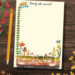 May include: A cream-colored notepad with a green vine border and the text "Beauty all around" at the top. The bottom of the notepad features a colorful illustration of a garden scene with flowers, mushrooms, worms, ants, and a skeleton.