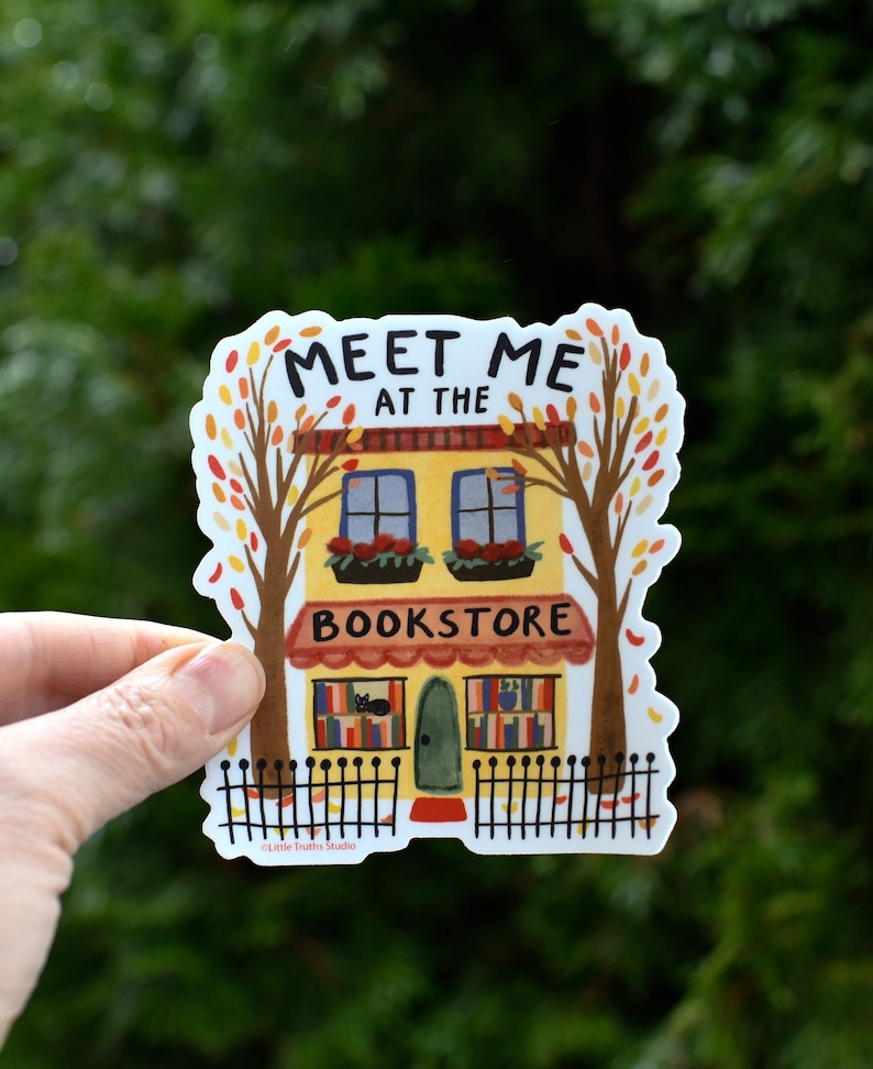 Cute Illustrated yellow bookstore vinyl sticker with autumn trees on each side and an iron fence in front that says meet me at the bookstore.