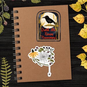 May include: Two stickers on a brown notebook. The first sticker features a black raven perched on a branch with a full moon behind it. The text reads "Quoth the Raven, Nevermore". The second sticker features a spider web with a spider hanging from a strand, a yellow flower, and blackberries.