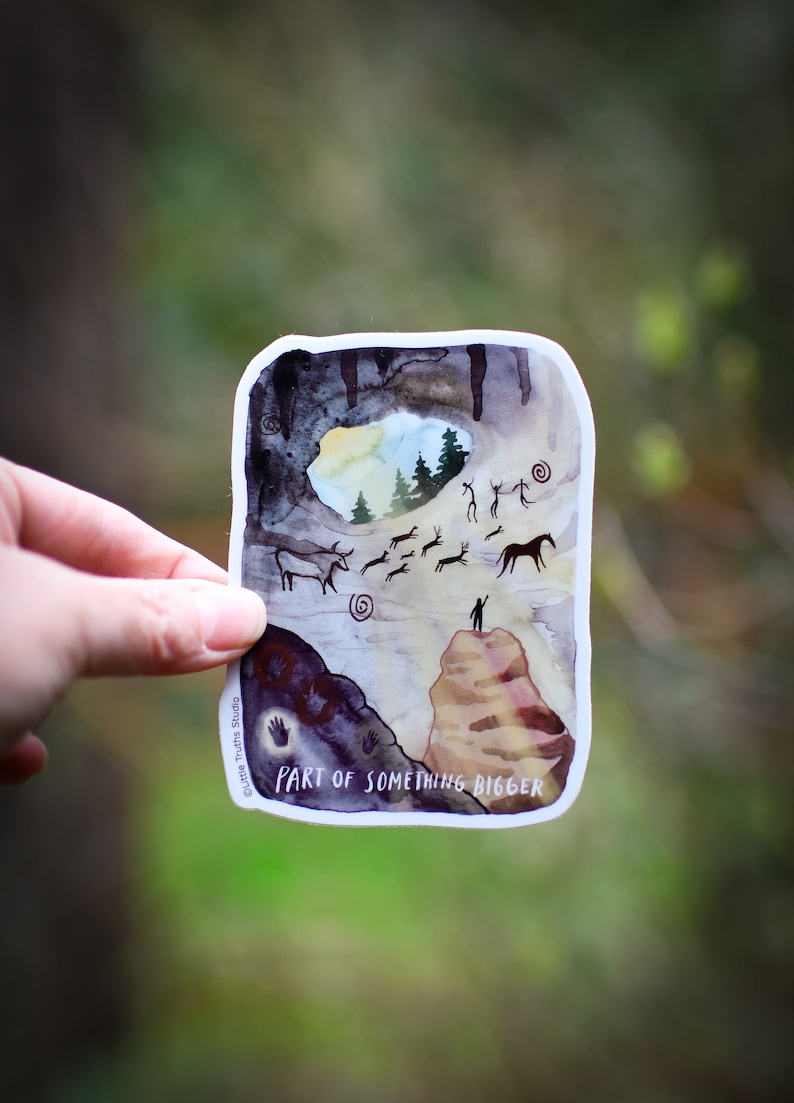 K&ouml;nnte beinhalten: Ein Aquarell-Aufkleber mit einer H&ouml;hlenszene, in der eine Person auf einer Klippe steht und auf einen Wald blickt. Die H&ouml;hle ist mit H&ouml;hlenmalereien von Tieren und Menschen gef&uuml;llt. Der Text "PART OF SOMETHING BIGGER" ist unten auf dem Aufkleber gedruckt.