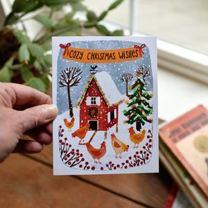May include: A Christmas card with a red barn scene, snow, and chickens. The card has the text "Cozy Christmas Wishes" on a banner. A green Christmas tree and bare trees are also in the winter scene.