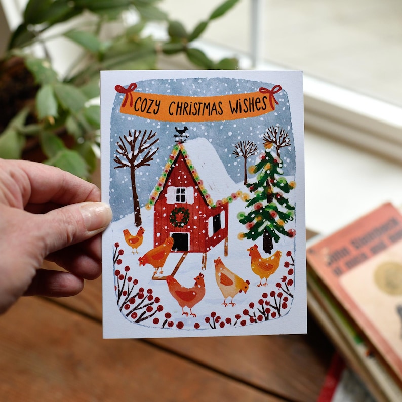 May include: A Christmas card with a red barn scene, snow, and chickens. The card has the text "Cozy Christmas Wishes" on a banner. A green Christmas tree and bare trees are also in the winter scene.