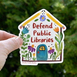 Puede incluir: Una pegatina blanca con un diseño en forma de casa. La casa tiene una puerta azul y lápices amarillos para el techo. El texto "Defend Public Libraries" está escrito en letras rojas. La pegatina está rodeada de flores y hojas.
