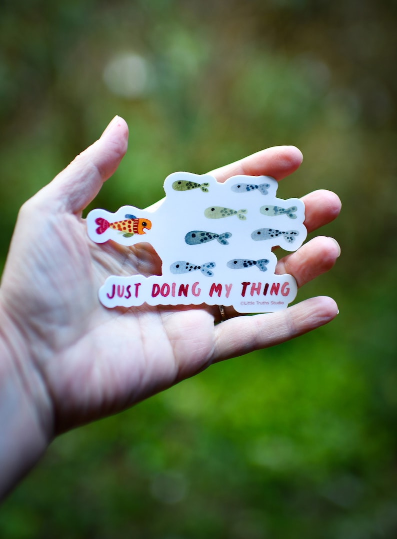 May include: A white sticker with a school of colorful fish and the text "Just doing my thing" in pink. The sticker is held in a hand.