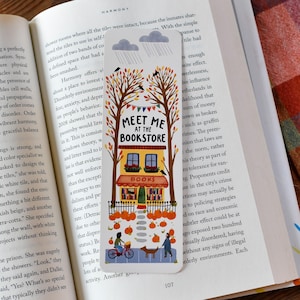 May include: A colorful bookmark with the text "Meet Me at the Bookstore." The illustration features a bookstore, trees, pumpkins, and figures. The bookmark is inserted into an open book.