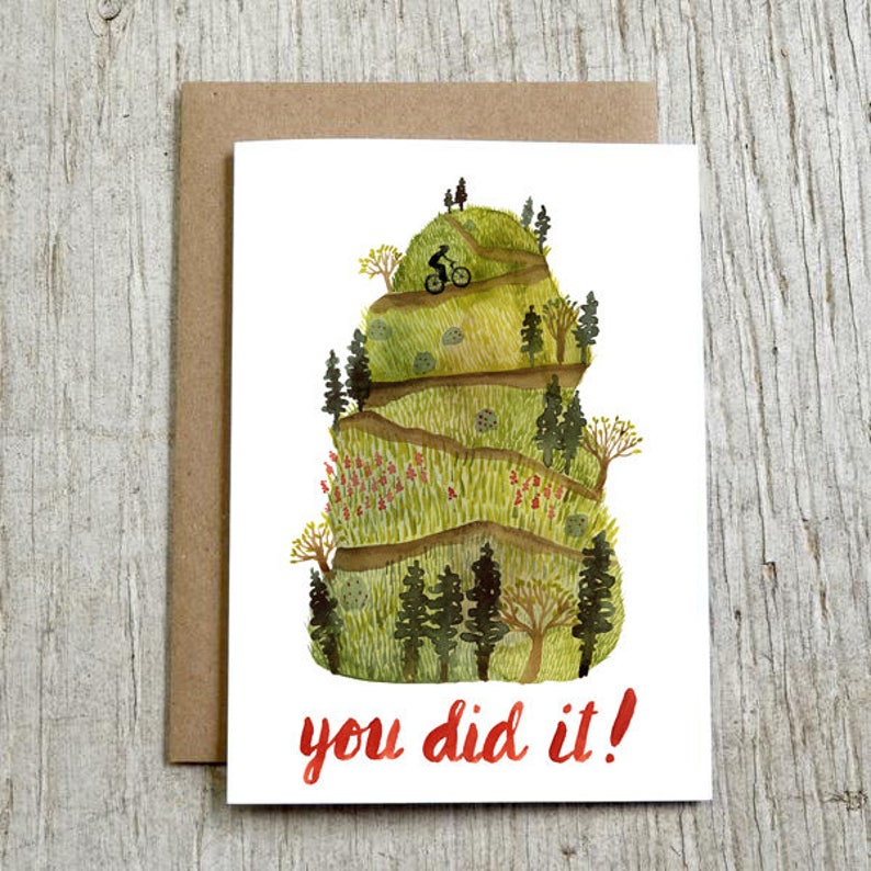May include: A watercolor illustration of a mountain with a winding path and a person riding a bicycle up the hill. The text "you did it!" is written in red at the bottom of the card.
