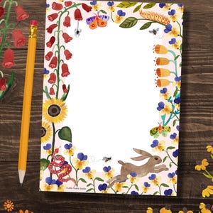 May include: A pink notepad with a white border and a floral design. The design includes red, yellow, blue, green, and purple flowers, a butterfly, a caterpillar, a snake, a rabbit, and a bee. The text "Little Truths Studio" is at the bottom of the notepad.