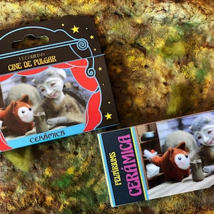 May include: Two rectangular product packages featuring the text FELTIRIUMS CINE DE PULGAR and CERÁMICA. The packaging displays a photographic illustration of a small brown felt fox and a grey felt figure interacting with a miniature ceramic object.