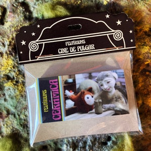 May include: A Feltiriums Cine de Pulgar pin in a clear plastic bag with a brown cardboard header. The header features white stars and the text Feltiriums Cine de Pulgar. The pin displays a scene with a felt fox and a figure working with clay.