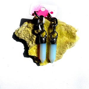 Pode incluir: Um par de brincos com pingentes em forma de cristal azul claro. Os brincos são suspensos por correntes e ganchos prateados. São exibidos em uma superfície amarela texturizada, com uma etiqueta branca com detalhes em rosa no fundo.