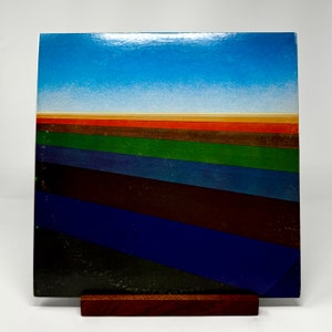 Puede incluir: Una portada de &aacute;lbum de vinilo vintage con un dise&ntilde;o abstracto colorido. La portada presenta un cielo azul con una l&iacute;nea de horizonte y una serie de rayas coloridas en tonos de rojo, naranja, marr&oacute;n, verde, azul y negro.
