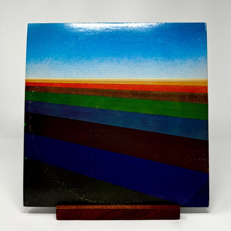 Puede incluir: Una portada de &aacute;lbum de vinilo vintage con un dise&ntilde;o abstracto colorido. La portada presenta un cielo azul con una l&iacute;nea de horizonte y una serie de rayas coloridas en tonos de rojo, naranja, marr&oacute;n, verde, azul y negro.