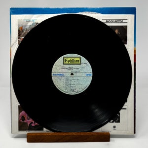 Puede incluir: Un disco de vinilo negro con una etiqueta amarilla y blanca. La etiqueta dice "Cotillion SD 5002, Tarkus, Emerson, Lake & Palmer, Atlantic". El disco est&aacute; en un soporte de madera.