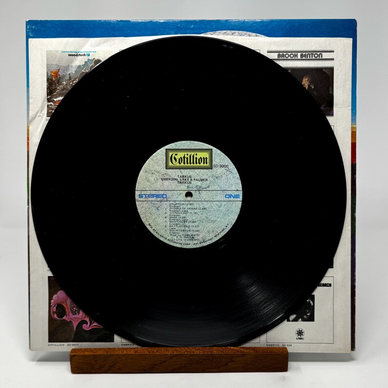 Puede incluir: Un disco de vinilo negro con una etiqueta amarilla y blanca. La etiqueta dice "Cotillion SD 5002, Tarkus, Emerson, Lake & Palmer, Atlantic". El disco est&aacute; en un soporte de madera.