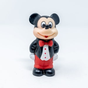 May include: A black and white Mickey Mouse figurine wearing a black tuxedo with a red bow tie and red pants. The figurine has a smiling face with black eyes and a red nose.