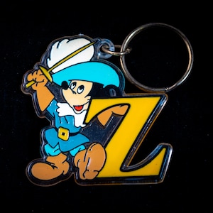 K&ouml;nnte beinhalten: Ein gelber Schl&uuml;sselanh&auml;nger aus Plastik mit einer Comicfigur, die ein Schwert h&auml;lt. Die Figur tr&auml;gt einen blauen Hut und ein wei&szlig;es Hemd. Der Schl&uuml;sselanh&auml;nger hat einen Metallring.