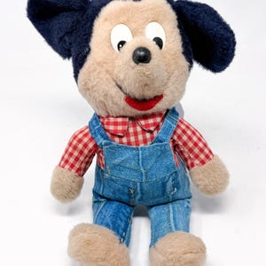Peut inclure: Une peluche vintage repr&eacute;sentant un personnage de dessin anim&eacute; de souris. Le jouet a un visage beige, des oreilles noires et porte une chemise &agrave; carreaux rouges et blancs et une salopette en jean bleu. La bouche est rouge et les yeux noirs.