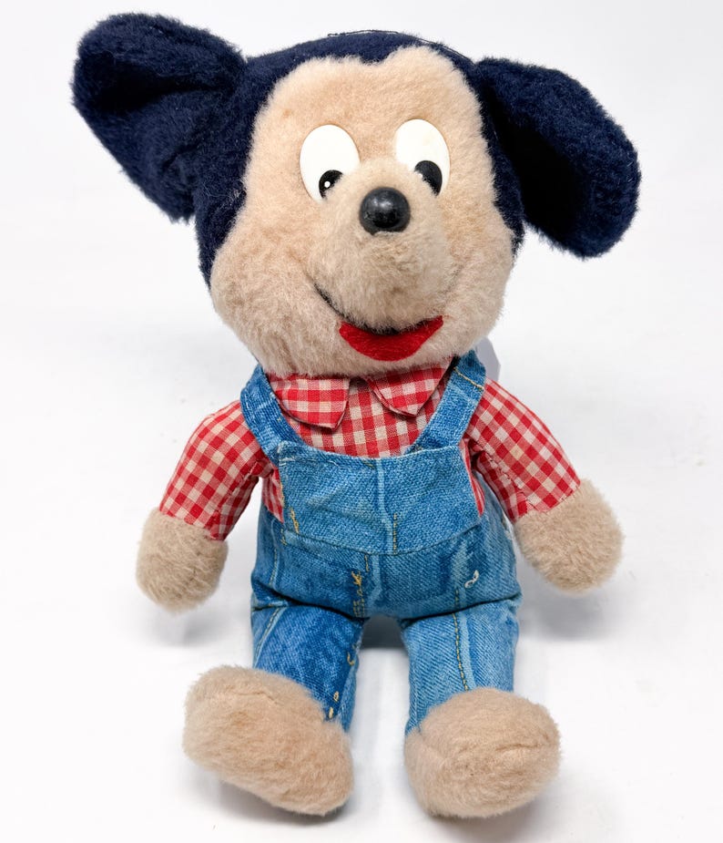 Peut inclure: Une peluche vintage repr&eacute;sentant un personnage de dessin anim&eacute; de souris. Le jouet a un visage beige, des oreilles noires et porte une chemise &agrave; carreaux rouges et blancs et une salopette en jean bleu. La bouche est rouge et les yeux noirs.