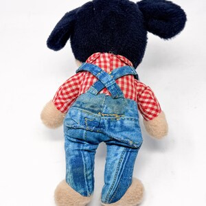 Peut inclure: Une peluche vintage de Mickey Mouse, vue de dos. Mickey porte une chemise &agrave; carreaux rouges et blancs, une salopette en jean bleue et a des oreilles noires. La peluche a des mains et des pieds beiges.