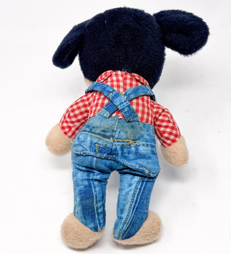 Peut inclure: Une peluche vintage de Mickey Mouse, vue de dos. Mickey porte une chemise &agrave; carreaux rouges et blancs, une salopette en jean bleue et a des oreilles noires. La peluche a des mains et des pieds beiges.