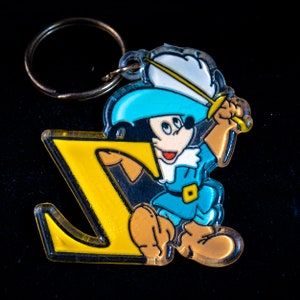 K&ouml;nnte beinhalten: Ein gelber Plastik-Schl&uuml;sselanh&auml;nger in Form des Buchstabens Z mit einer Comicfigur, die ein Schwert h&auml;lt. Die Figur tr&auml;gt einen blauen Hut, ein blau-wei&szlig;es Hemd und braune Stiefel.