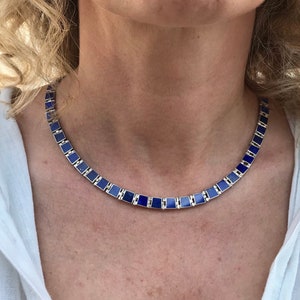 May include: A silver-toned necklace with rectangular, deep blue gemstone inlays. The necklace is worn around the neck and is set against a white shirt. The gemstones are a rich, dark blue color.