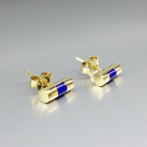Pu&ograve; includere: Un paio di orecchini a bottone color oro. Ogni orecchino ha un design cilindrico con una vivace striscia di smalto blu al centro. Gli orecchini sono presentati su una superficie grigio chiaro.