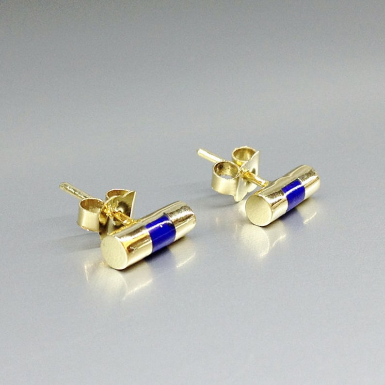 Pu&ograve; includere: Un paio di orecchini a bottone color oro. Ogni orecchino ha un design cilindrico con una vivace striscia di smalto blu al centro. Gli orecchini sono presentati su una superficie grigio chiaro.