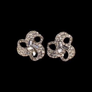 May include: A pair of silver-toned, clip-on earrings. Each earring features a swirling, ribbon-like design encrusted with numerous small, clear crystals. Rectangular crystals are set in the center of each earring. The background is black.